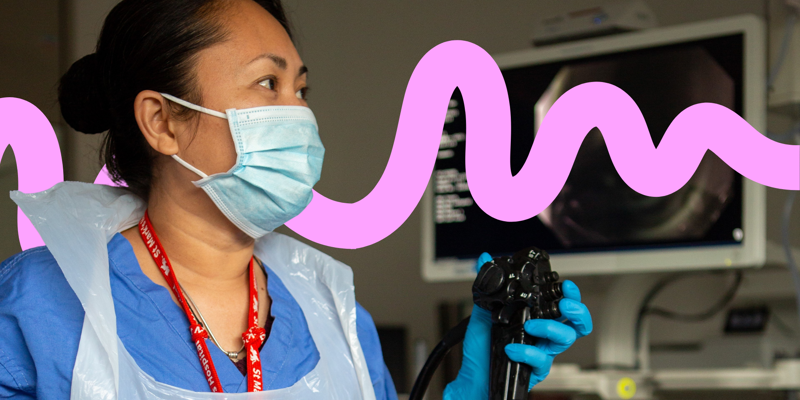 A healthcare worker in blue scrubs and a face mask holds an endoscope, with a medical monitor displaying an internal view in the blurred background.