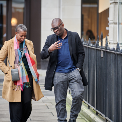 Black man in dark jeans with blue top and black coat and and woman in dark outfit with camel coat and multi-colour scarf walking through london street talking