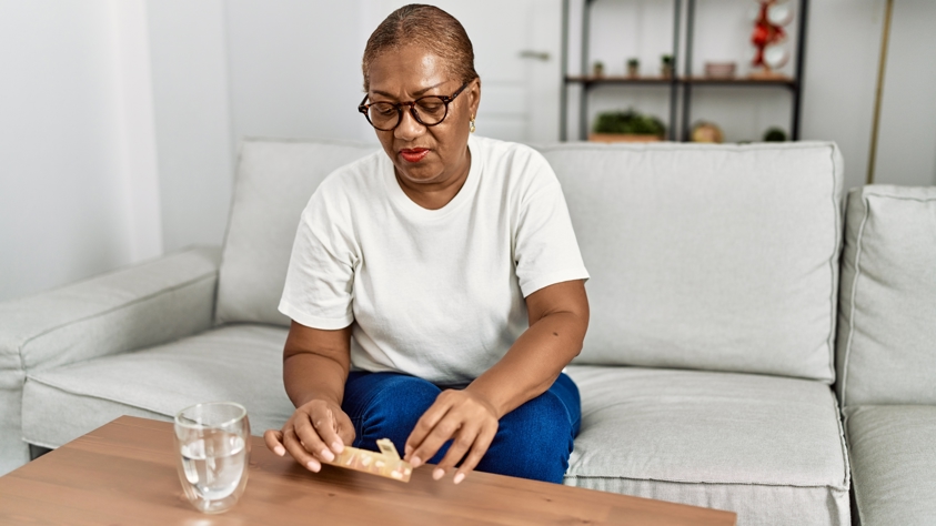 An older black female is taking a tablet out of a tablet organiser. She has a glass of water ready