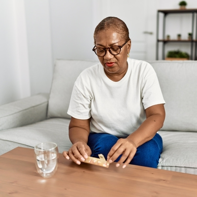 An older black female is taking a tablet out of a tablet organiser. She has a glass of water ready