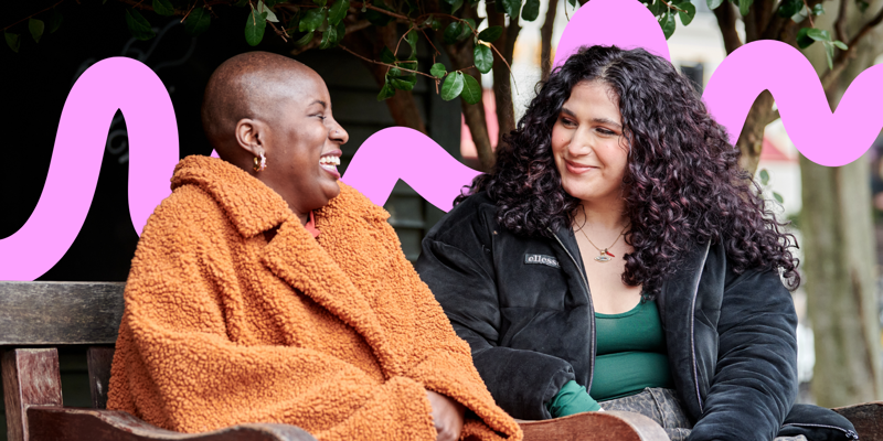 The image shows two women seated closely on a wooden bench. The woman on the left has a shaved head and is laughing, wearing a bright orange, fluffy coat that contrasts with her dark skin. She has hoop earrings and is seated with her hands on her lap. The woman on the right has long, curly hair with hints of red and is wearing a black, padded jacket over a green top. She appears to be listening intently with a gentle smile. Both are surrounded by lush greenery, creating a warm and inviting atmosphere. 
