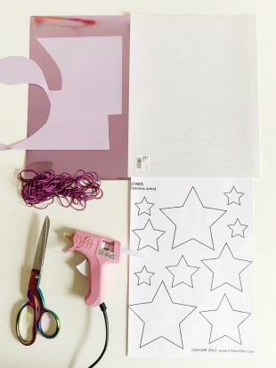 Craft equipment. Scissors, glue-gun, white glitter felt sheet, purple ribbon and paper with stars drawn on.
