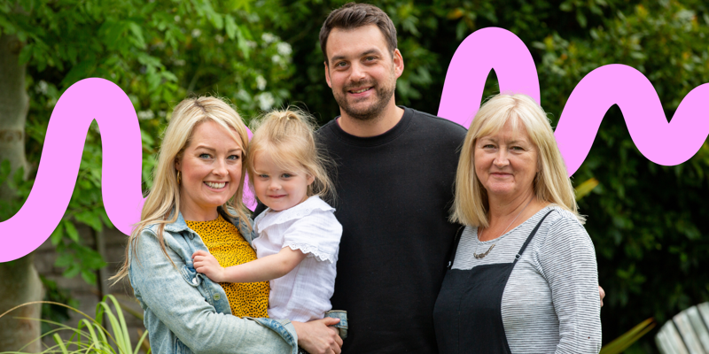 A family of four, including a young girl held by a woman, a man, and an older woman, poses outdoors against a backdrop of green foliage and pink art.